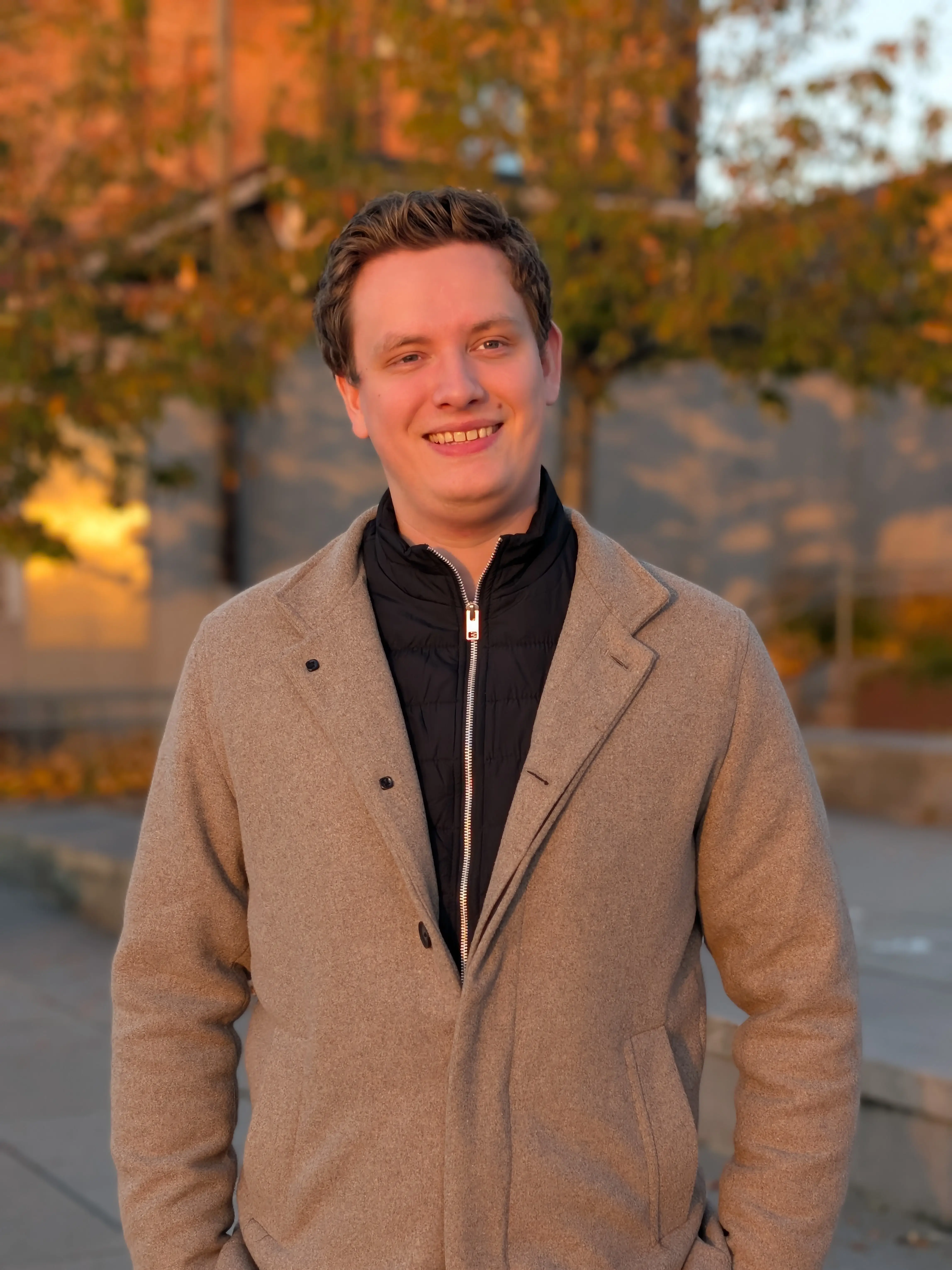 Smiling young man wearing a beige coat and black zip-up jacket standing outdoors in warm evening light.