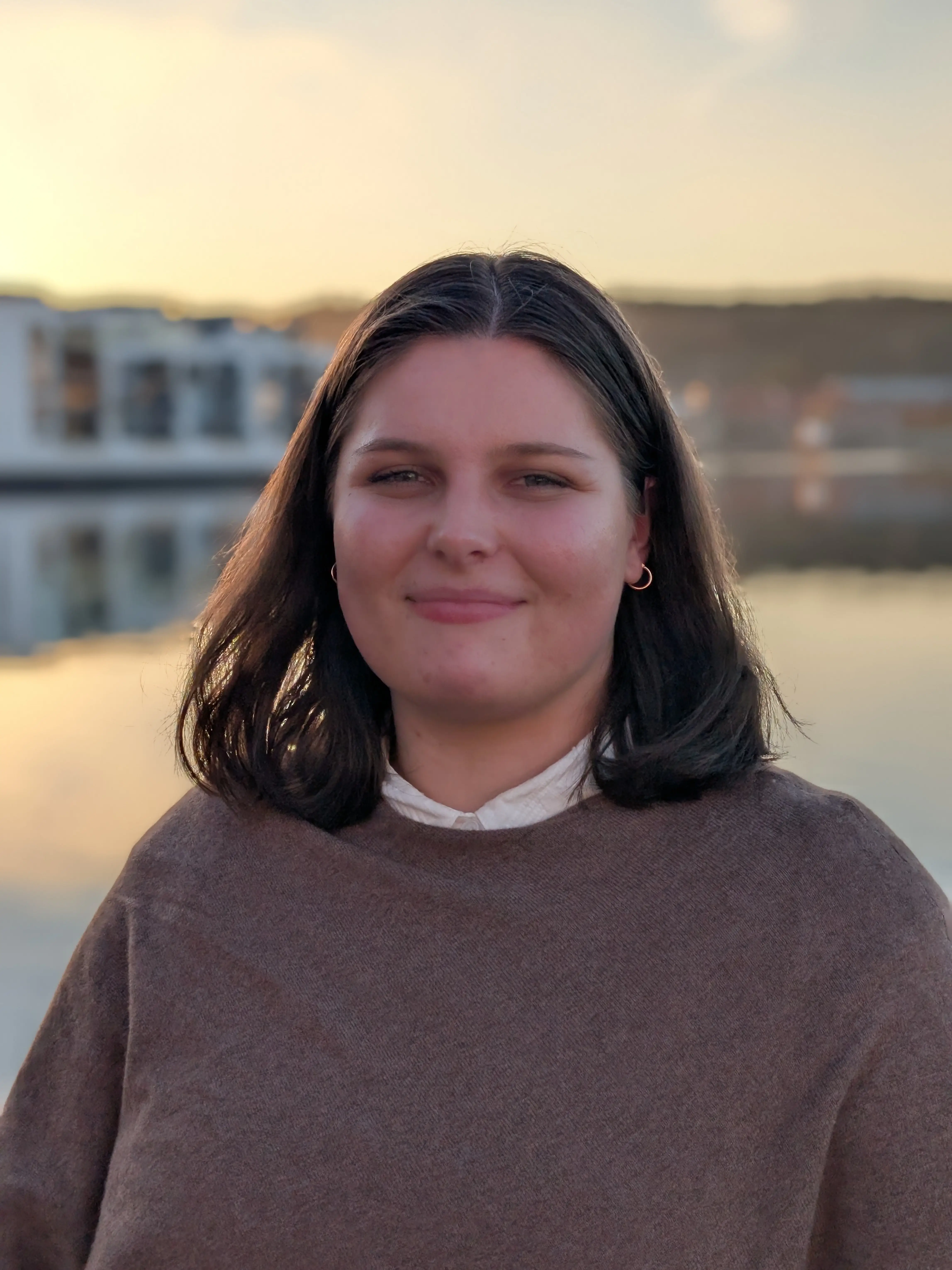 Smiling woman with dark hair wearing a brown sweater standing outdoors near a body of water at sunset.