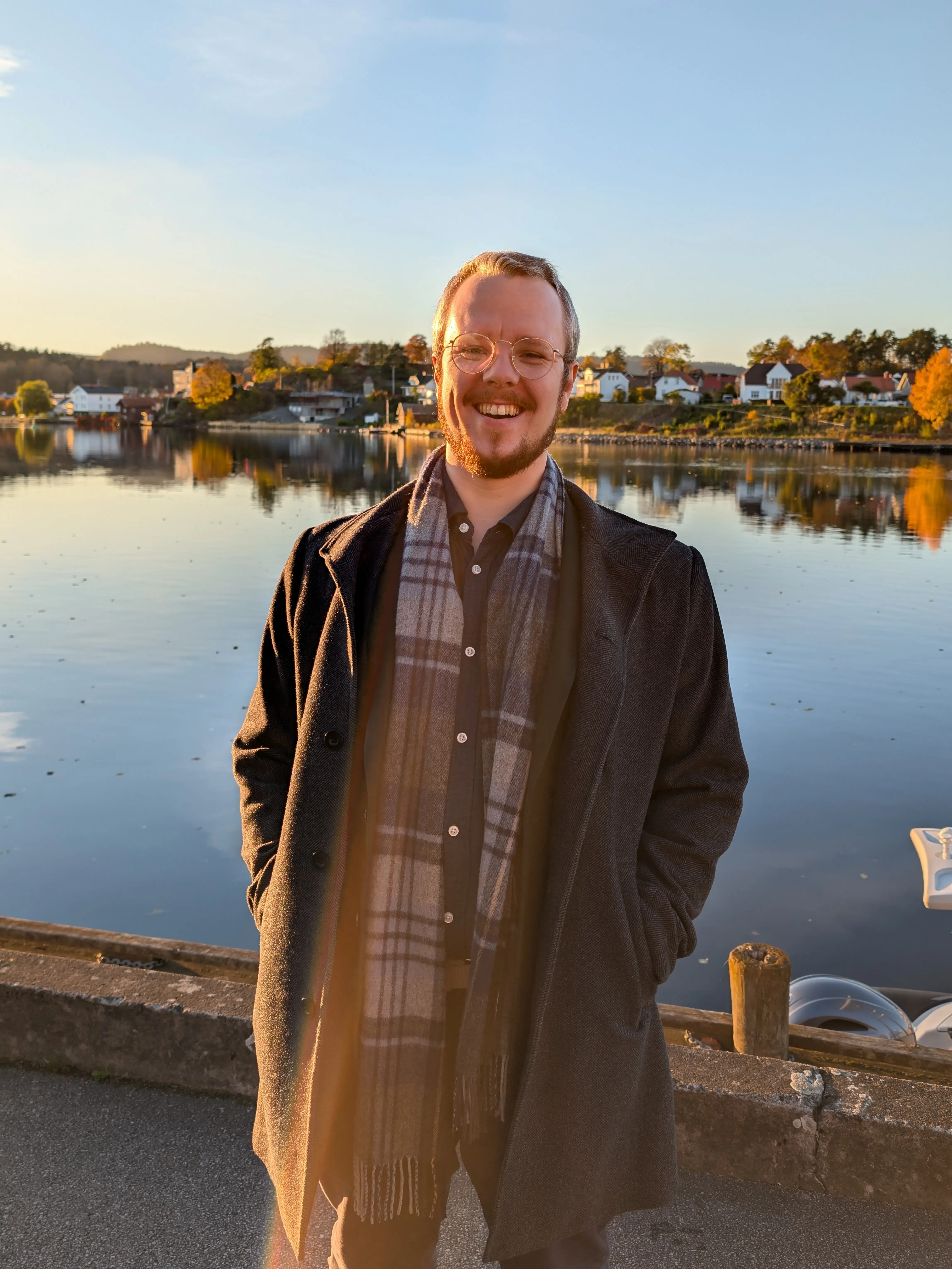 Smiling man in glasses and a dark coat with a plaid scarf standing by a calm waterfront during sunset.